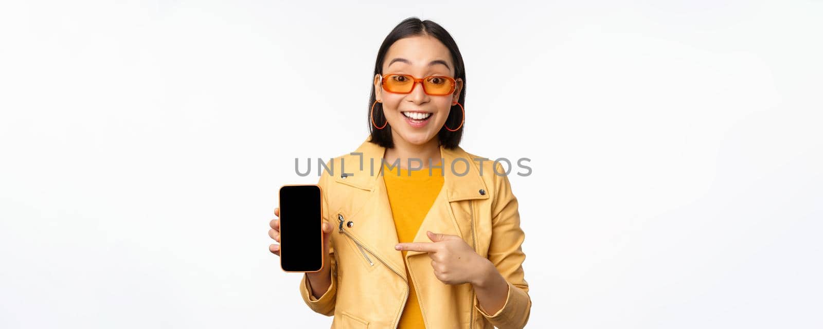 Portrait of stylish korean girl in sunglasses, smiling, pointing finger at smartphone screen, showing mobile phone application, standing over white background.