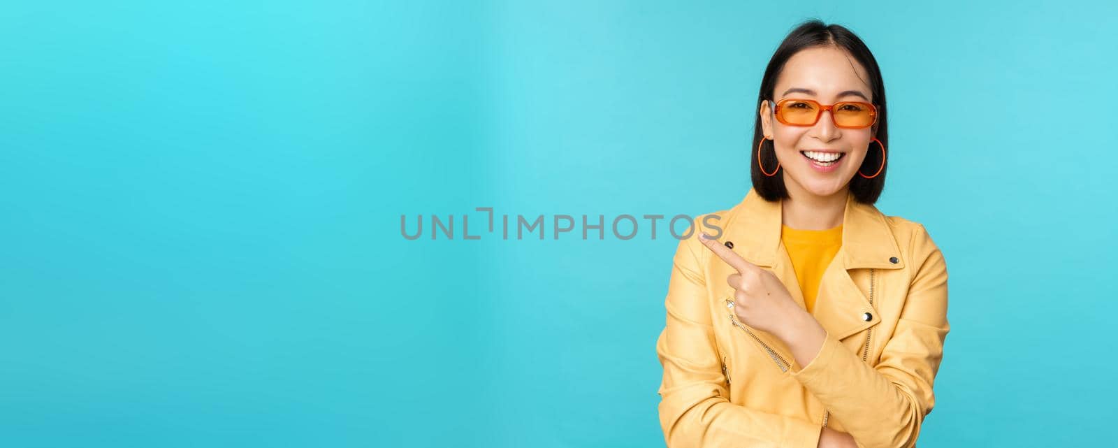 Smiling asian woman in sunglasses, pointing finger left, showing banner, logo or advertisement, standing over blue background. Copy space