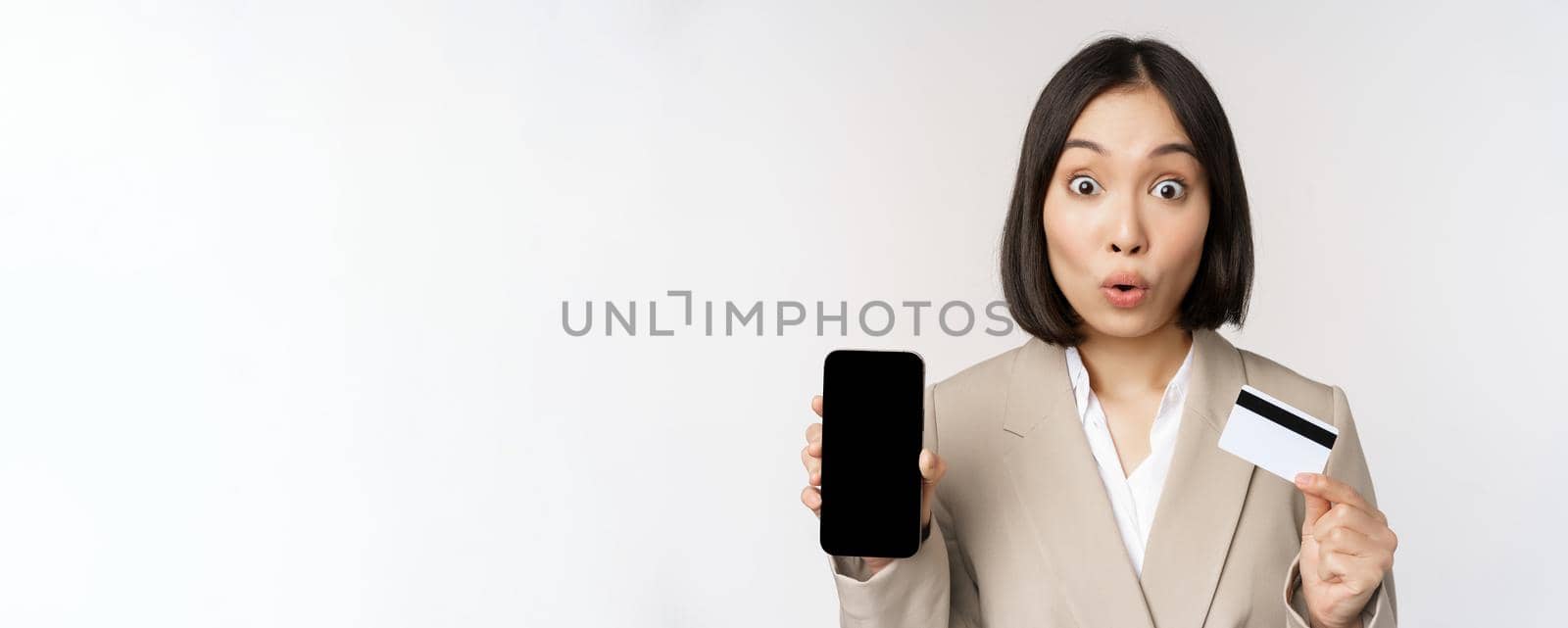 Corporate woman with happy, enthusiastic face, showing credit card and smartphone app screen, standing in suit over white background by Benzoix