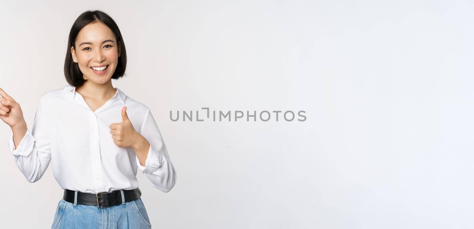 Image of young asian business woman, smiling while pointing finger left and showing thumbs up, recommending product, praise, standing over white background.