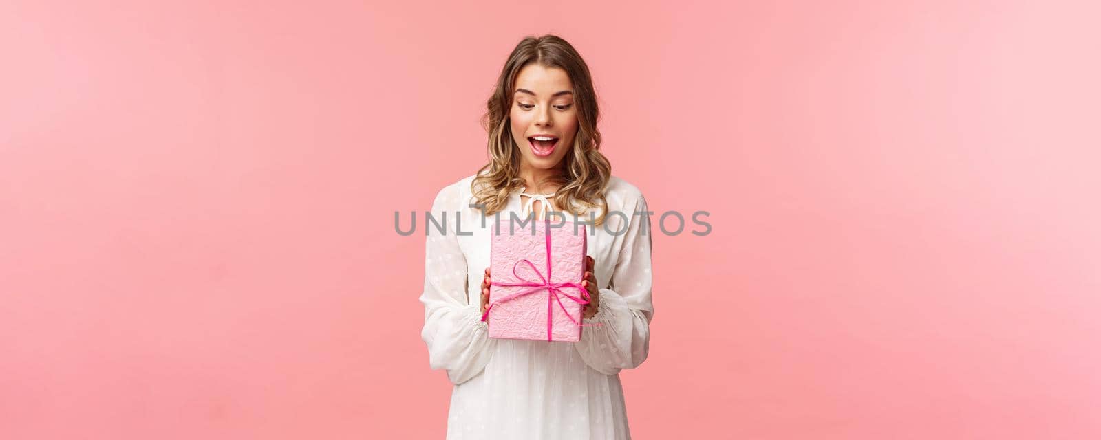 Holidays, celebration and women concept. Portrait of surprised charming young blond girl receive surprise gift, holding present in pink box look at it amused, curious whats inside, studio background.