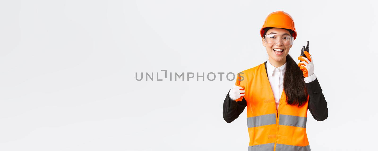 Satisfied happy smiling asian female engineer, industrial technician in safety helmet and uniform showing thumbs-up while praising great work using walkie-talkie, give permission to work by Benzoix