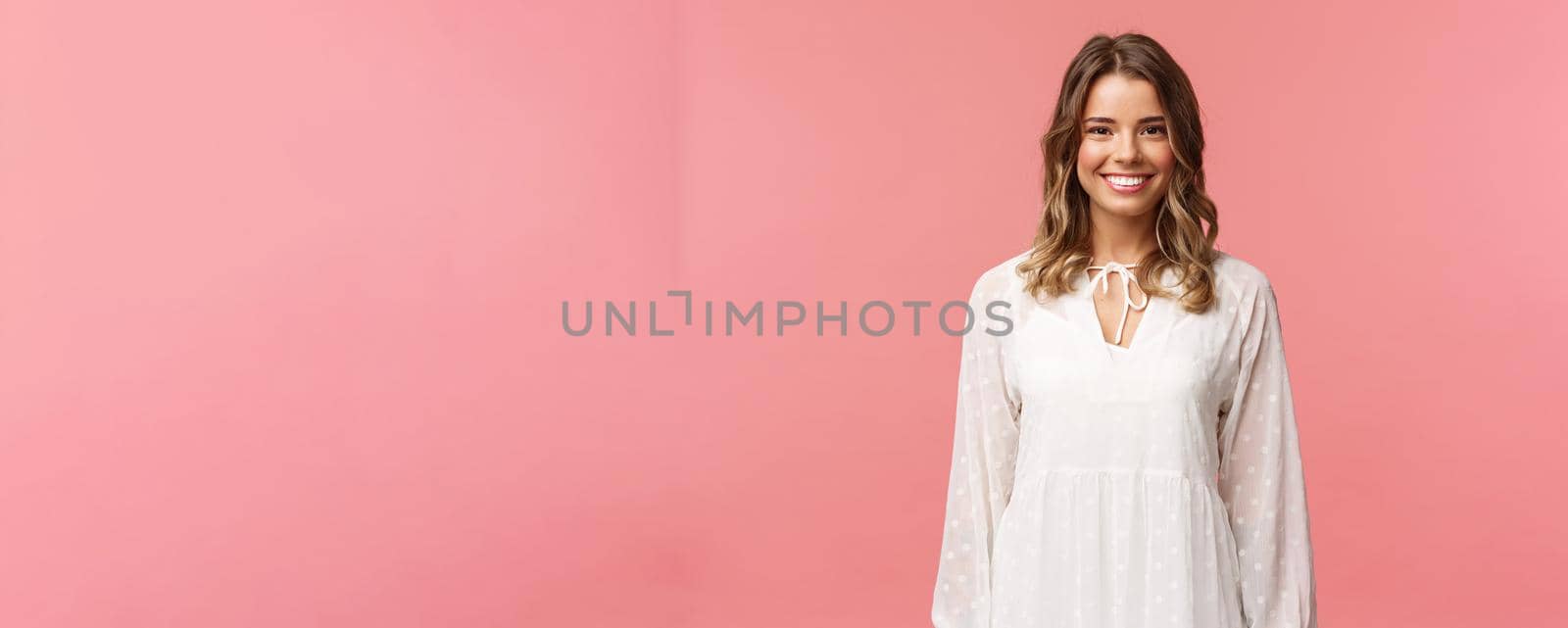 Spring, beauty and women concept. Portrait of tender feminine blonde girl with white beaming smile standing in light cute dress over pink background with happy attitude, positive emotions.