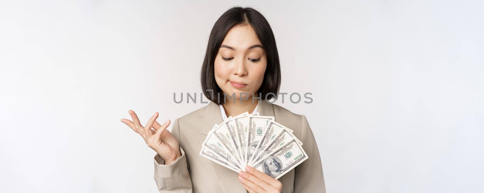 Image of asian corporate woman, happy businesswoman showing money, cash dollars and thinking, standing in suit over white background.