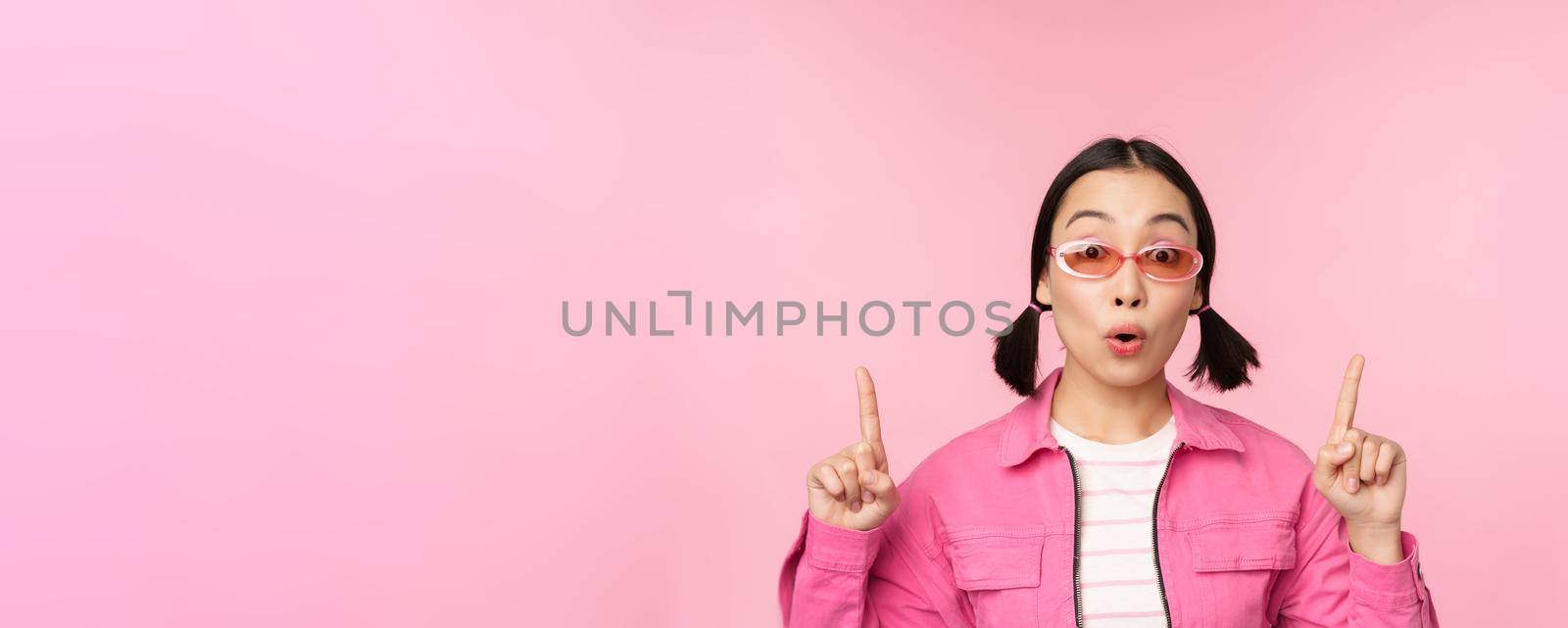 Close up portrait of asian woman gasping, looking surprised, pointing finger at banner, advertisement, standing over pink background.