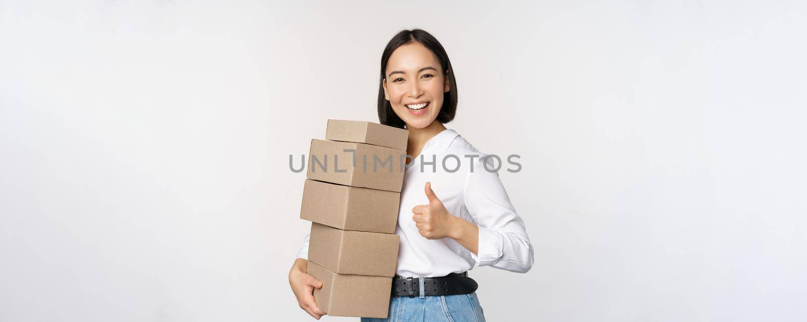 Image of happy modern asian woman showing thumbs up, holding boxes delivery goods, standing against white background.