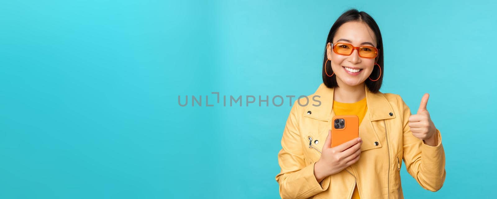 Smiling asian woman showing thumbs up, recording on mobile phone, using smartphone app and recommending it, standing over blue background.