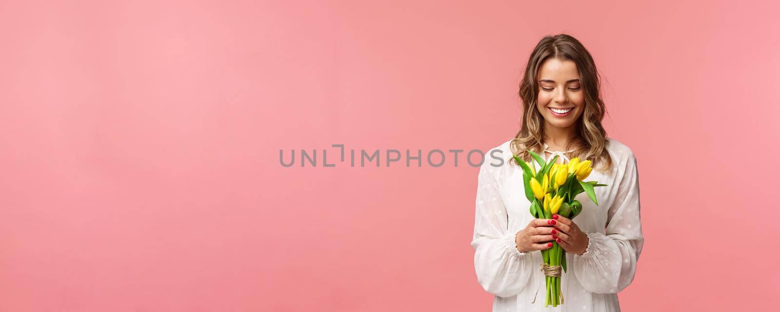 Holidays, beauty and spring concept. Portrait of lovely, romantic blonde girl in white dress, holding yellow tulips, looking at beautiful flowers, smiling and blushing, pink background.