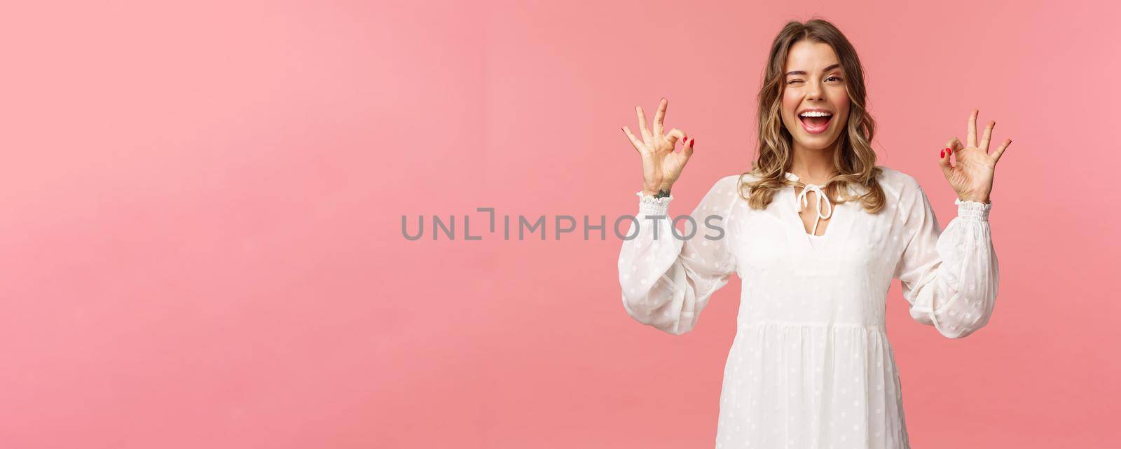 Beauty, fashion and women concept. Portrait of carefree unbothered blond feminine woman in white dress, guarantee quality, wink say no problem, smiling pleased, rate good product, pink background.