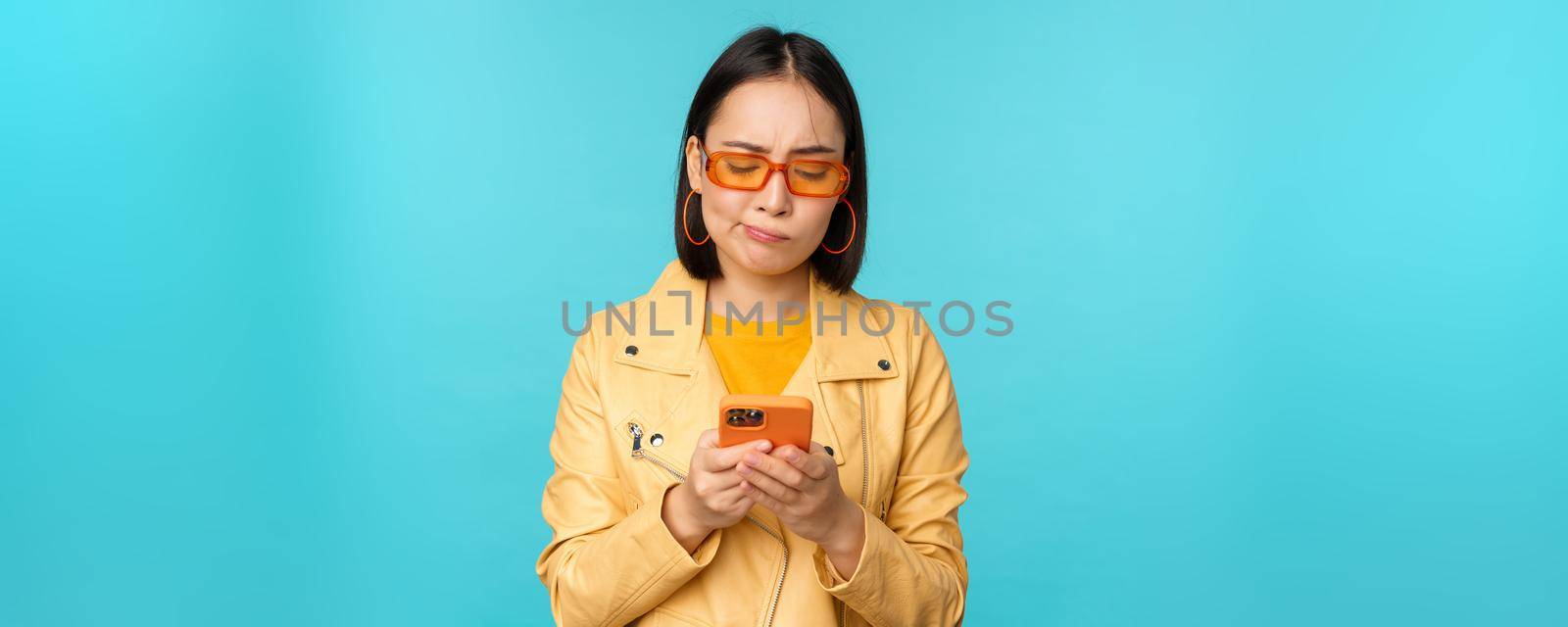Image of chinese young woman looks disappointed at her smartphone, looking at mobile phone with regret, standing over blue background.