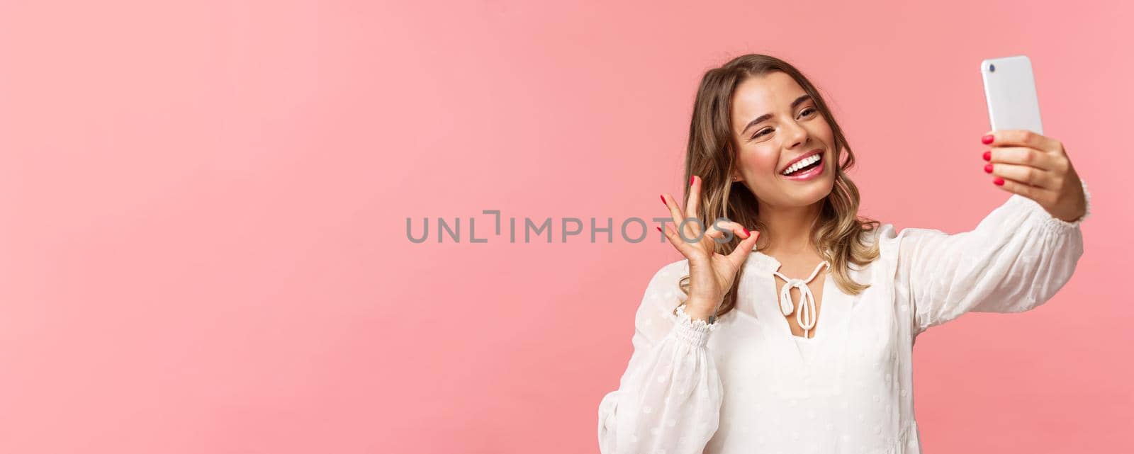 Close-up of satisfied good-looking blond girl in white dress, taking selfie, record mobile phone video, show okay satisfactory sign with pleased nod, smiling agree or recommend, pink background.