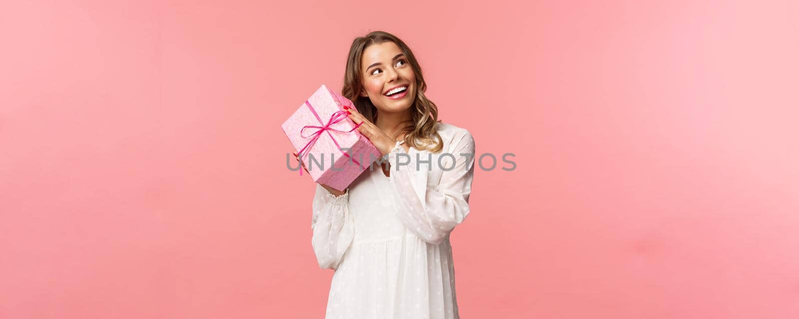 Holidays, celebration and women concept. Portrait of curious attractive young blond girl celebrating birthday, shaking pink gift box to found out whats inside, smiling intrigued look camera.