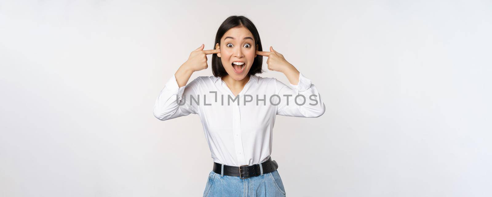 Young annoyed asian girl close ears and screaming, fed up with loud noise, terrible music, standing over white background by Benzoix