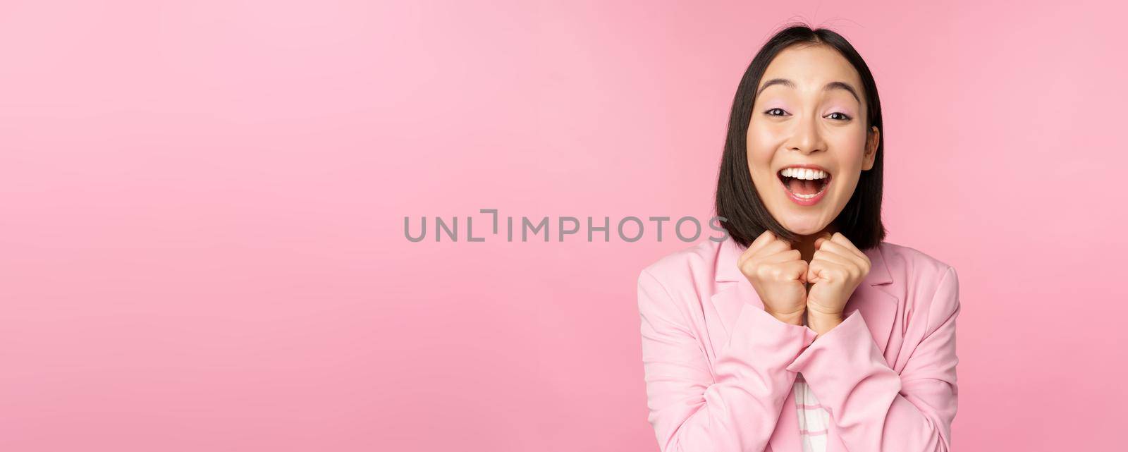 Portrait of happy asian office lady, ceo businesswoman in suit rejoicing and laughing, winning, celebrating, achieve goal and rejoice, standing over pink background.