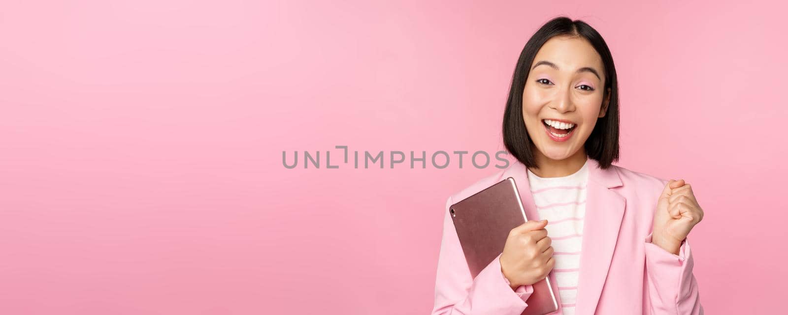 Enthusiastic asian businesswoman with digital tablet, screaming with joy, triumphing, standing over pink background in suit. Copy space