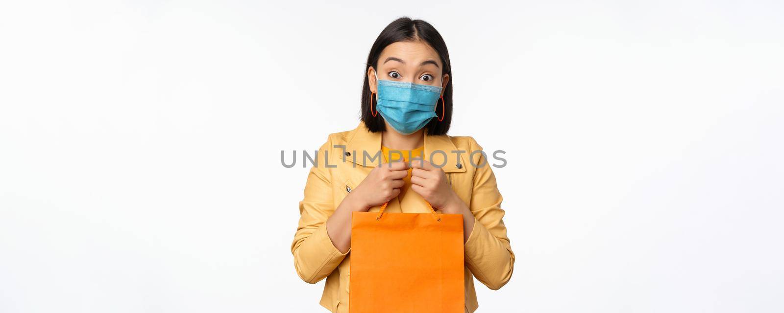 Covid-19 and shop concept. Young asian stylish woman, wearing medical face mask, holding shopping bag, going in malls during pandemic, white background.