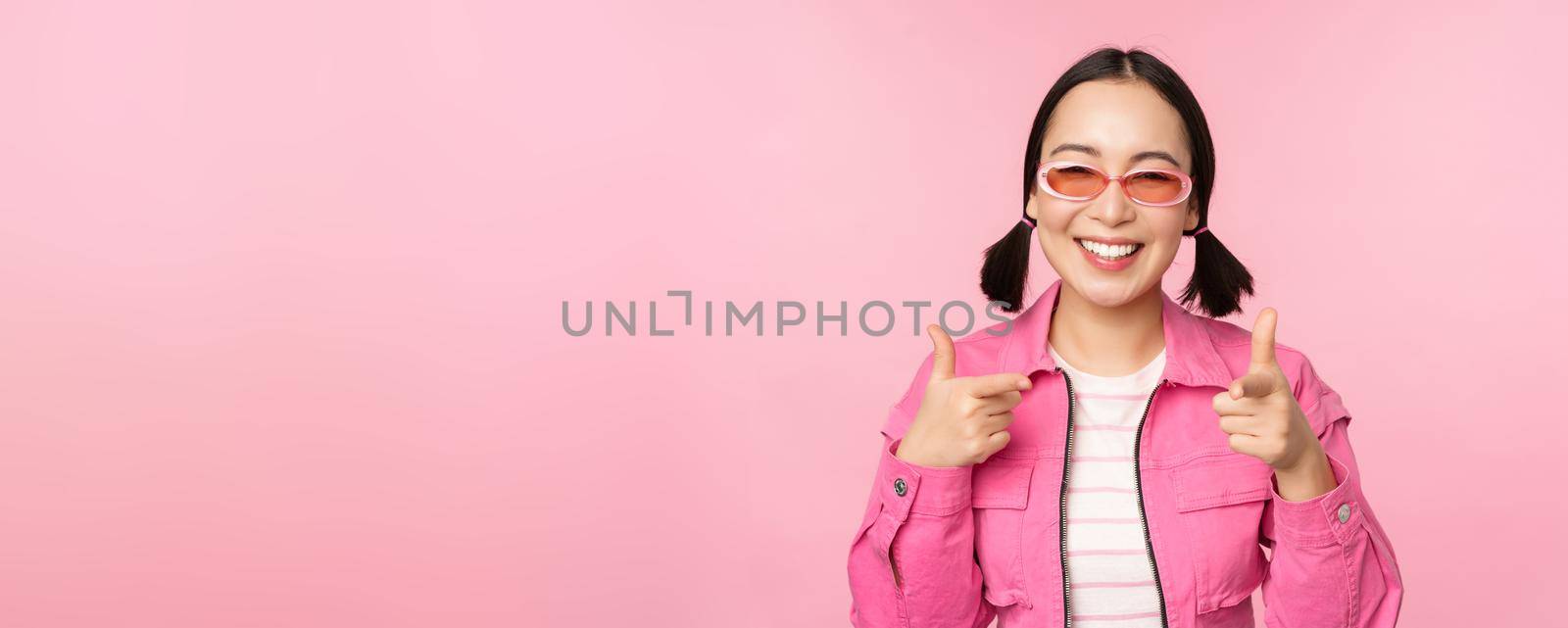 Close up portrait of modern asian girl in sunglasses smiling, pointing fingers at camera, praise you, inviting or complimenting, standing over pink background by Benzoix
