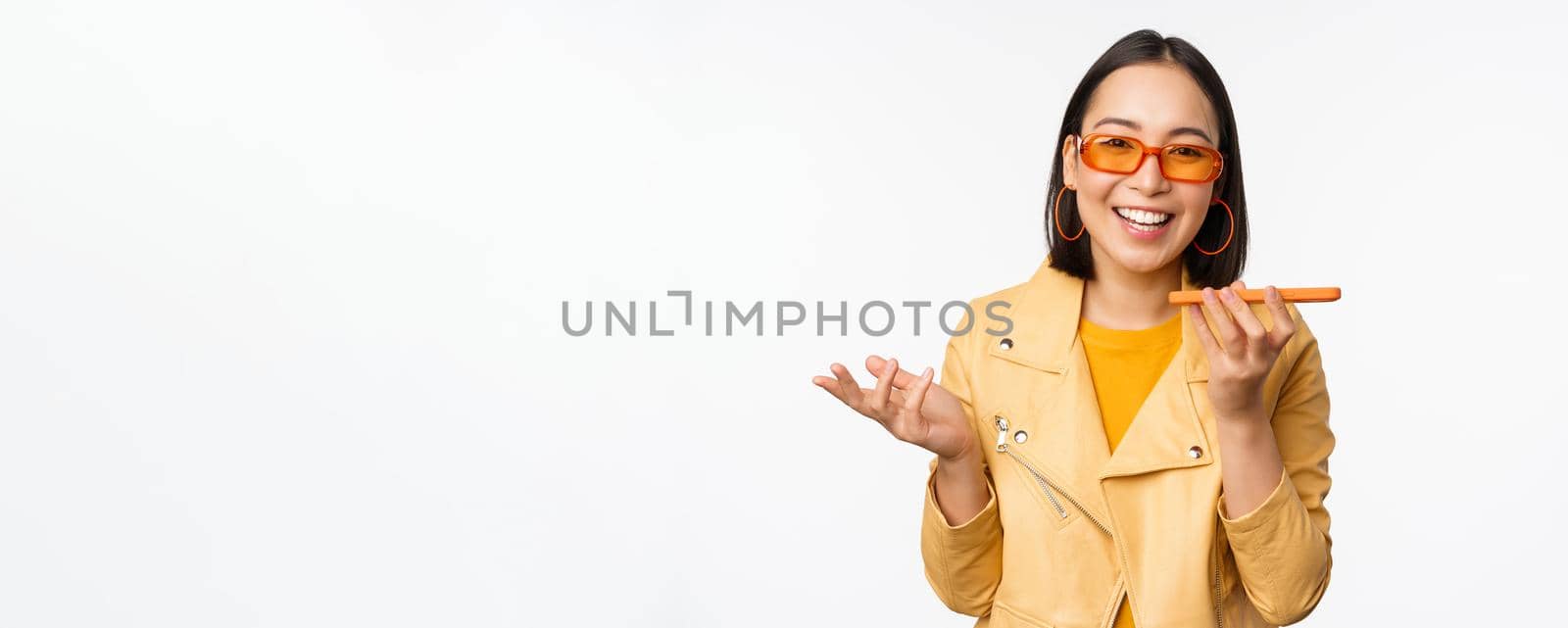 Image of happy asian girl talking on speakerphone, recording, translating her voice with mobile phone app, talking in smartphone dynamic, standing over white background by Benzoix