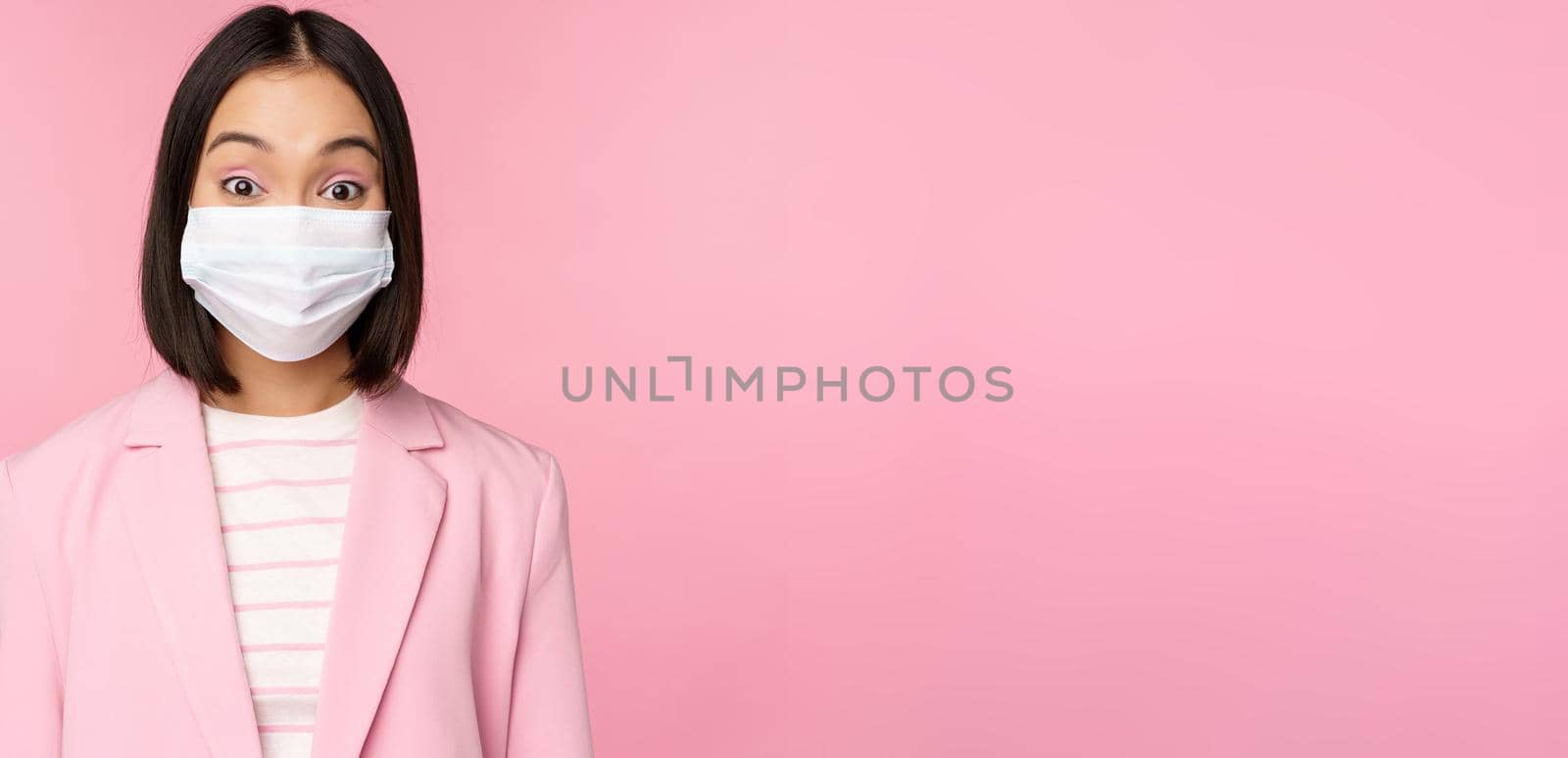 Portrait of asian businesswoman in medical face mask, wearing suit, concept of office work during covid-19 pandemic, standing over pink background by Benzoix