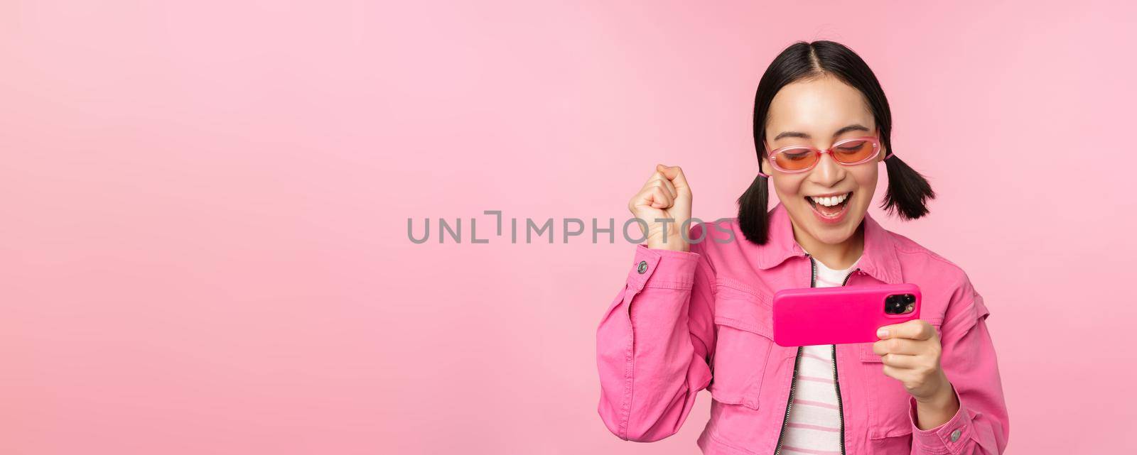 Happy smiling korean girl winning on mobile phone, looking at horizontal smartphone screen and rejoicing, achieve goal, celebrating, standing over pink background by Benzoix