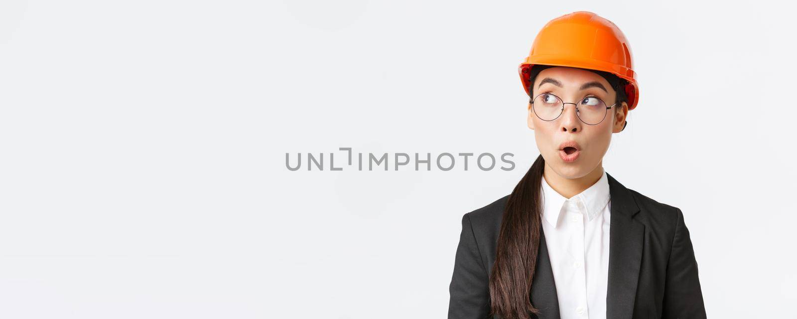 Close-up of impressed asian female chief architect, industrial engineer in safety helmet and business suit, looking left surprised and amazed, standing white background at factory.