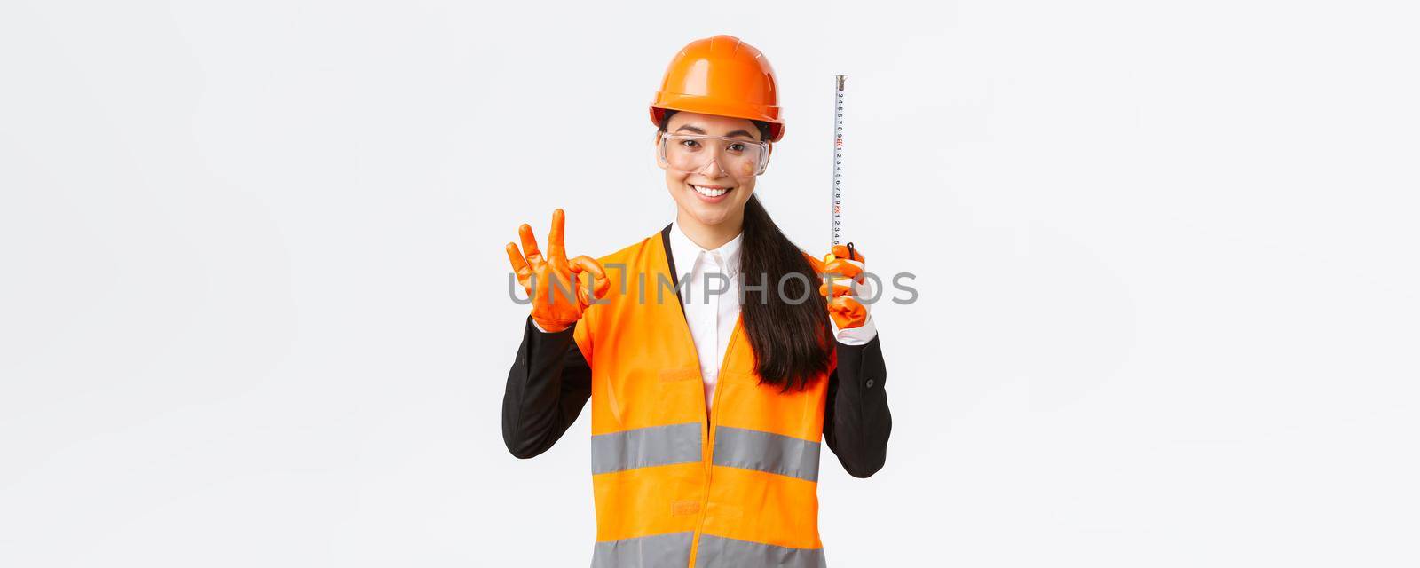 Satisfied asian female construction engineer, architect or inspector at enterprise showing okay gesture and tape measure, smiling pleased, give permission, approve measurements are valid by Benzoix