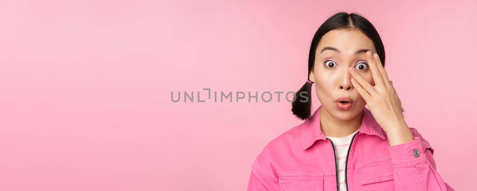 Close up portrait of young asian girl looking surprised, express amazement and wonder, peeking through fingers, standing over pink background.