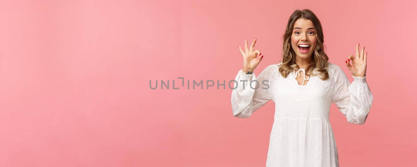 Portrait of happy and pleased good-looking blond woman in white dress, recommend great product, leave positive review, show okay signs and smiling satisfied, standing pink background.