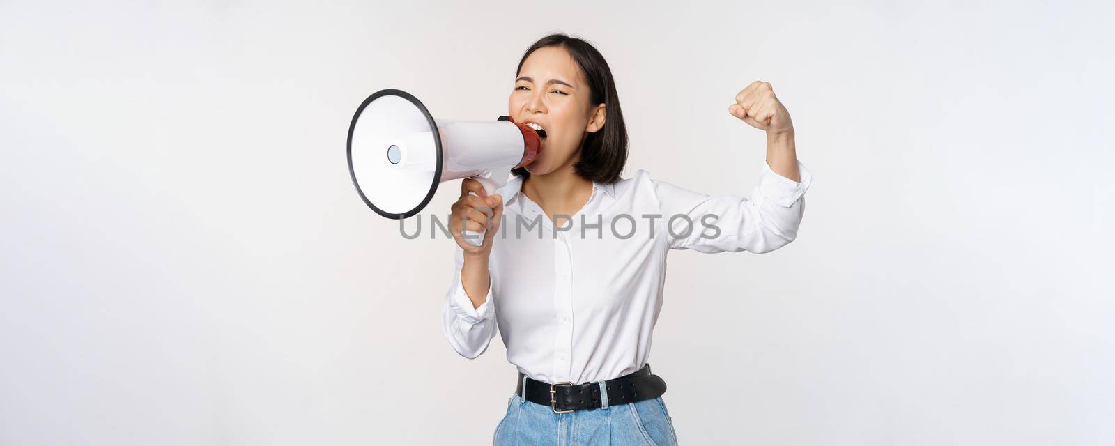 Enthusiastic asian woman, girl activist shouting at protest, using megaphone, looking confident, talking in loudspeaker, protesting, standing over white background.