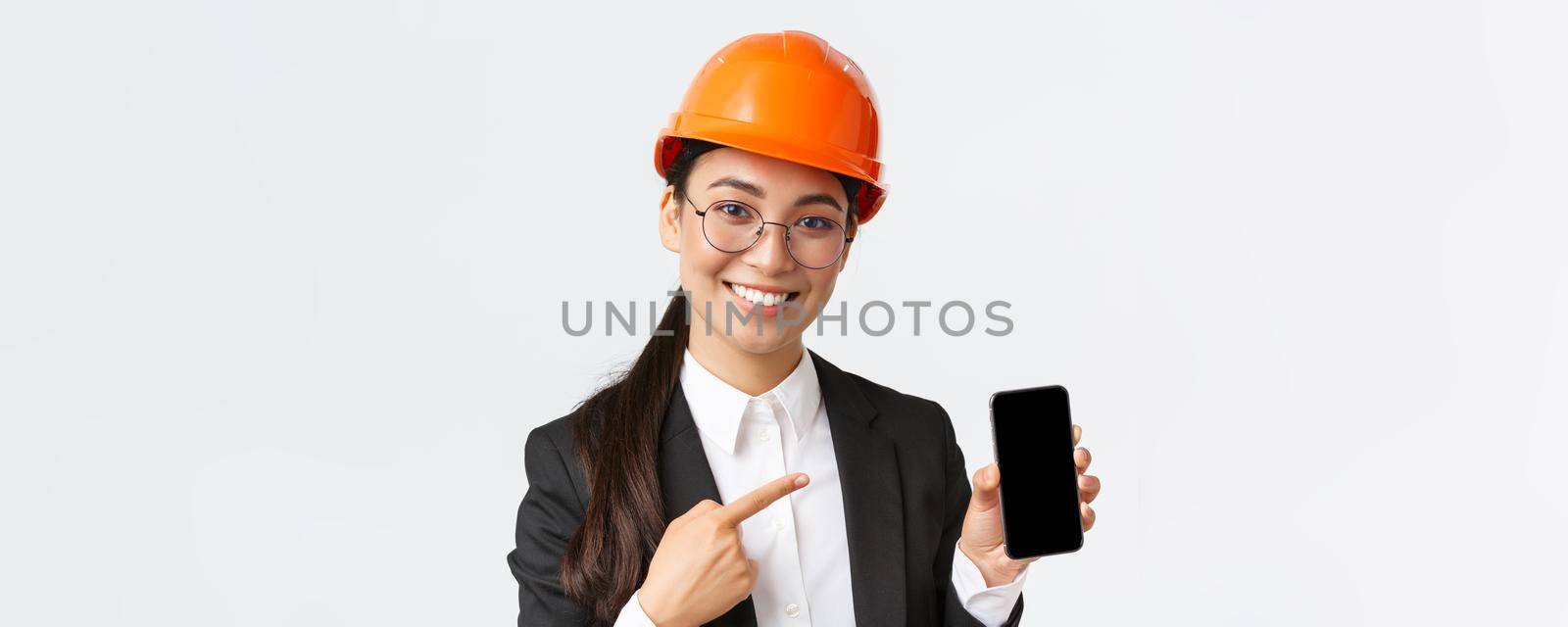 Close-up of smiling professional female asian construction engineer, architect in suit and safety mask introduce application, pointing finger at smartphone display, white background by Benzoix