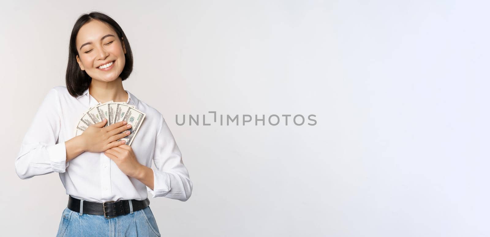 Happy asian woman hugging money dollars and smiling satisfied, standing over white background. Copy space