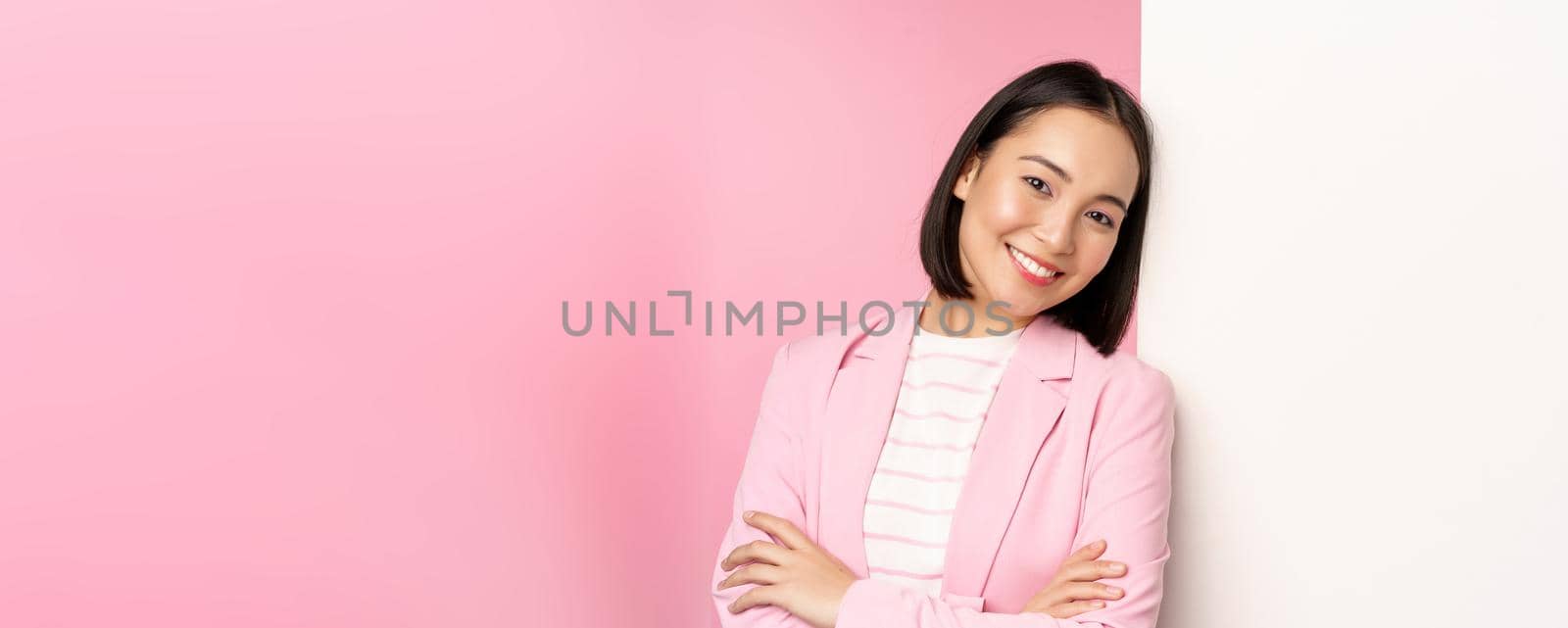 Confident, successful japanese office lady in suit, cross arms, looking as professional at camera, leaning on white wall with advertisement, empty copy space for logo, pink background by Benzoix