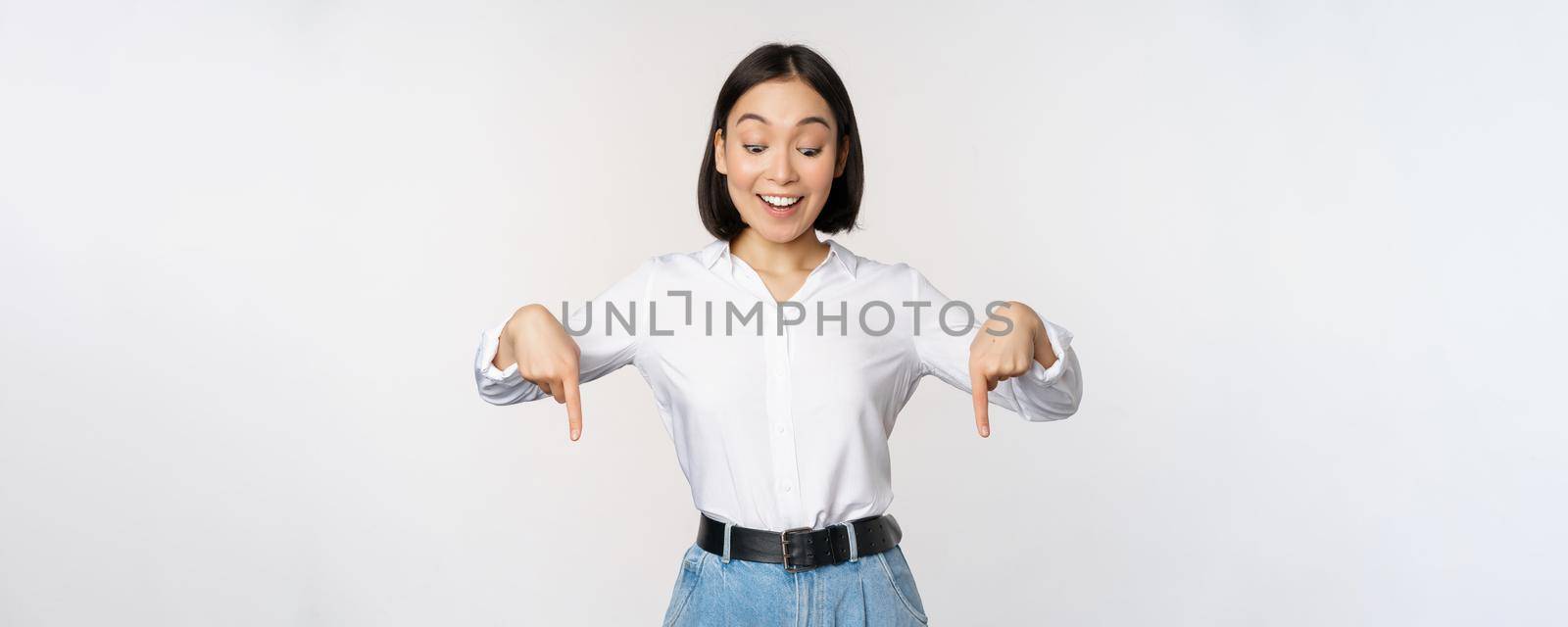 Portrait of happy asian woman pointing fingers down and looking below at advertisement, showing info banner, advertising, standing over white background.