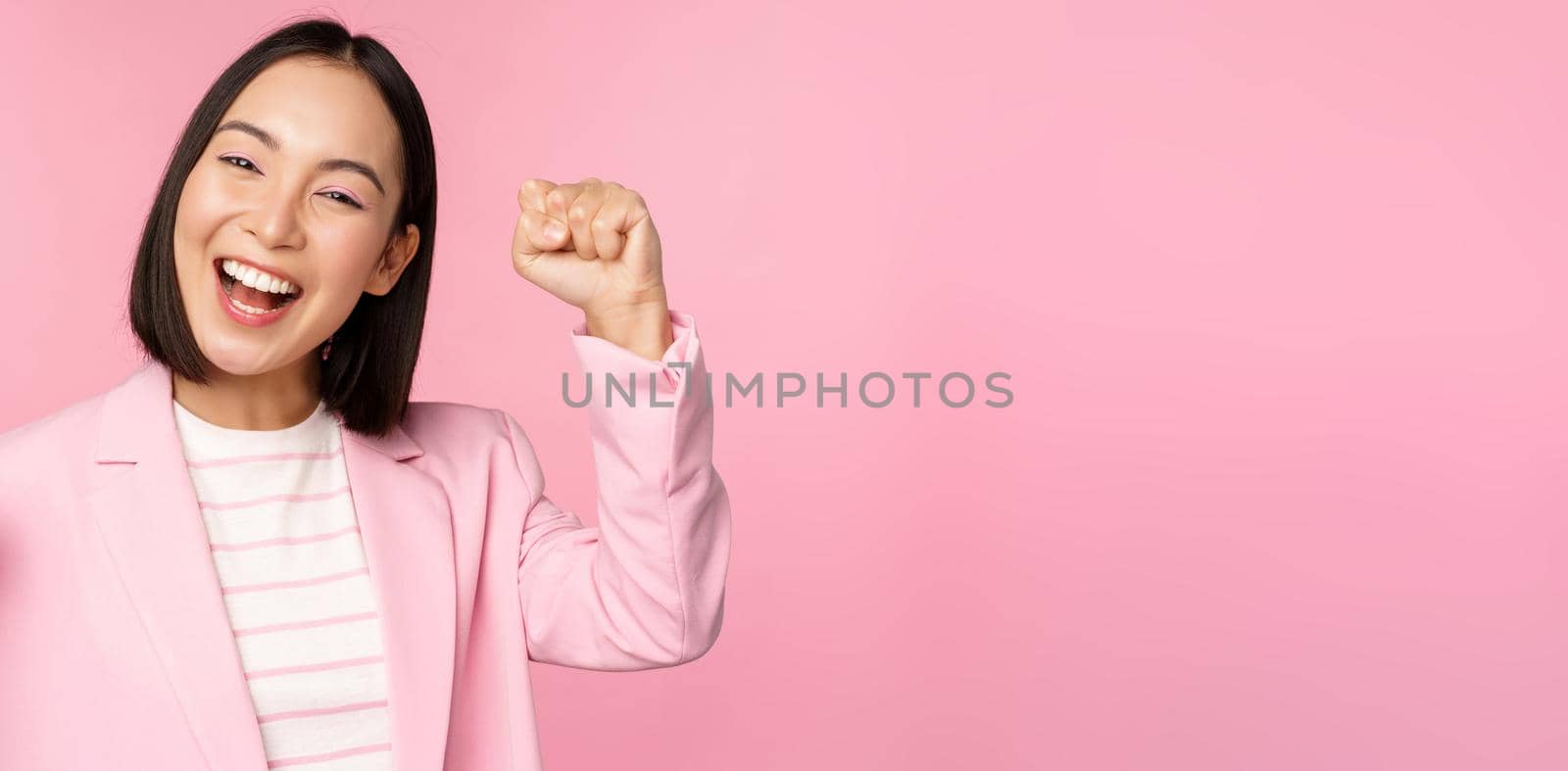 Enthusiastic asian corporate woman, businesswoman raising hand up and cheering, triumphing, winning and celebrating, standing over pink background by Benzoix