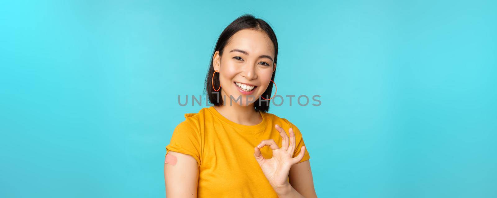 Vaccine campaign from covid-19. Happy healthy asian girl showing okay sign and smiling, bandaid on shoulder from coronavirus vaccination, blue background by Benzoix