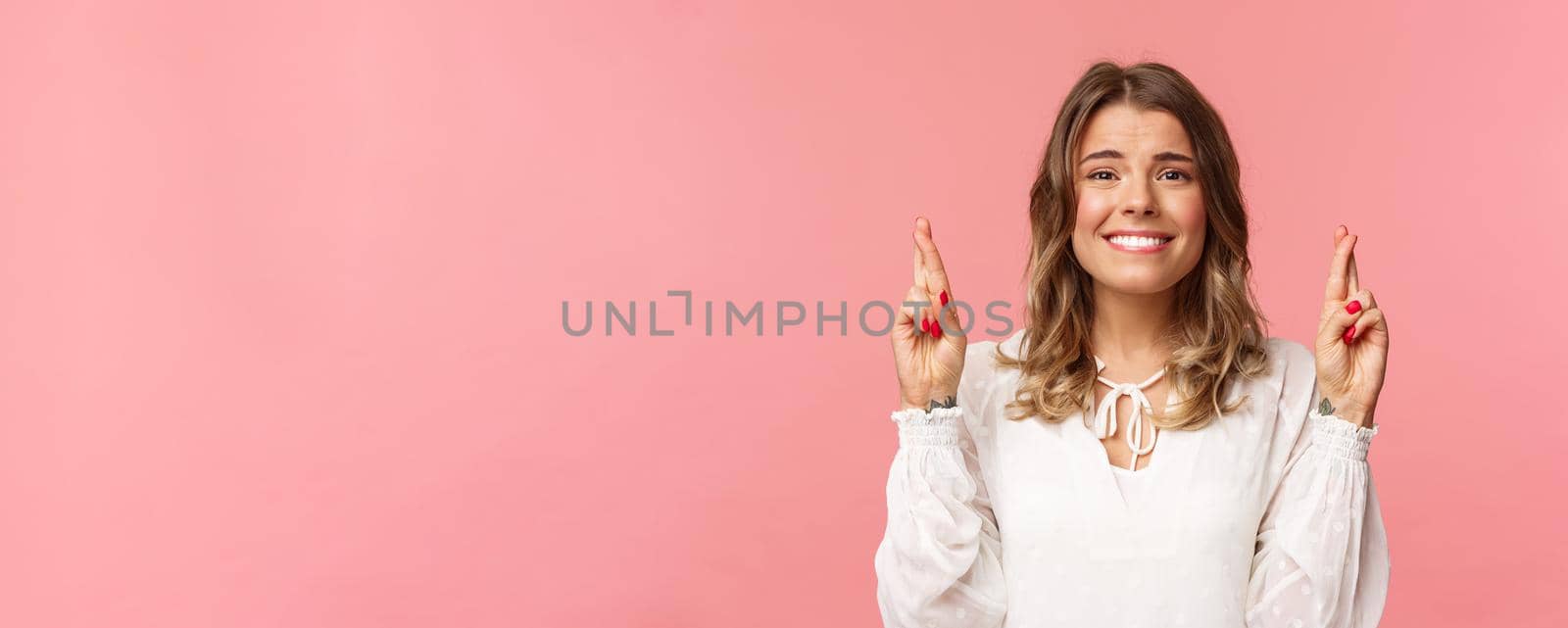 Close-up portrait of hopeful beautiful blond girl making wish, anticipating important results, cross fingers good luck and smiling as feeling slightly nervous, wear white dress, pink background.