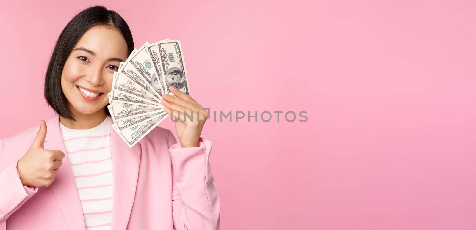 Microcredit, investment and business people concept. Young asian businesswoman, corporate lady showing money, cash dollars, thumbs up, recommending company, pink background by Benzoix