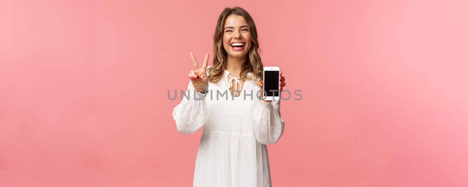 Portrait of kawaii optimistic and happy young girl in white dress, show mobile phone display and peace sign, laughing feeling cheerful and glad to share awesome app, useful link, pink background.