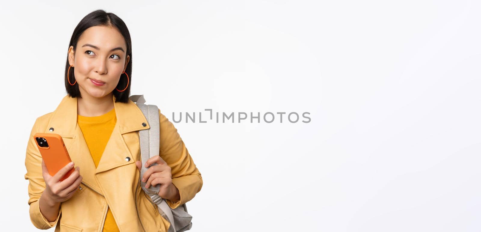 Asian girl traveller with backpack, holding mobile phone, using smartphone app, looking thoughtful, standing over white background.