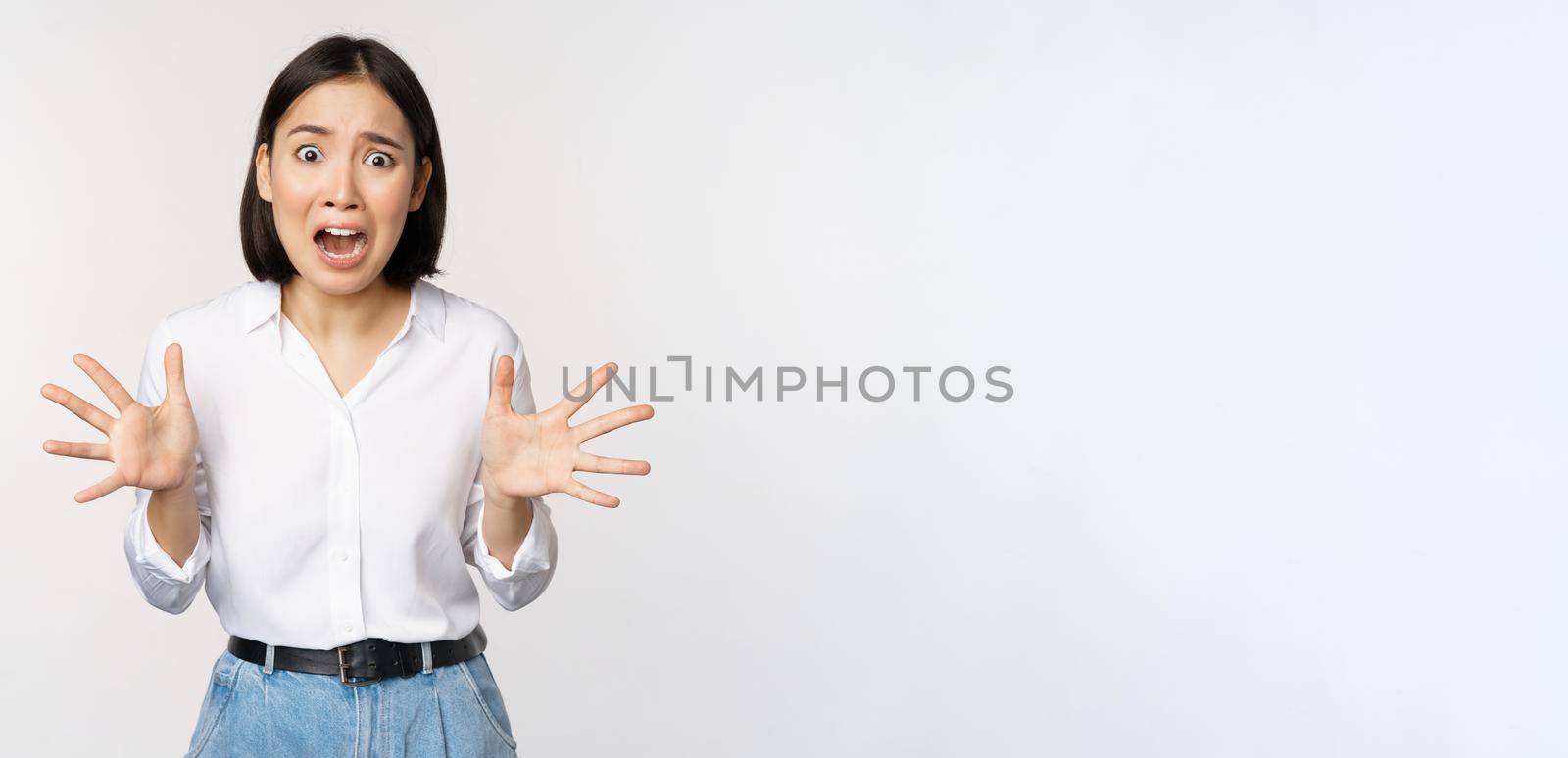 Asian woman looks at camera and screams in panic. Young korean girl looking anxious, panicking, shaking hands and shouting, standing over white background.