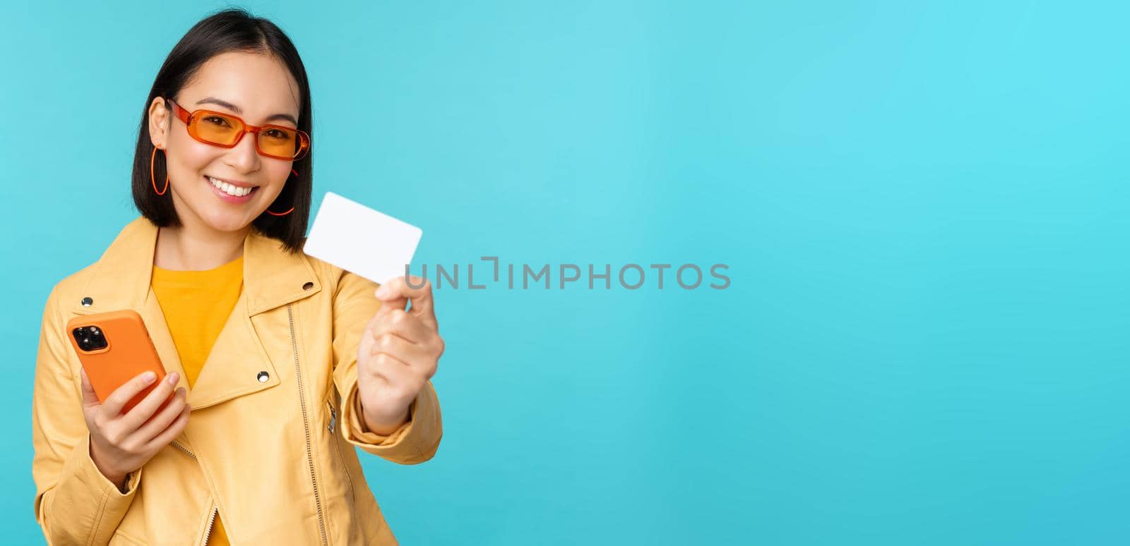 Online shopping. Stylish young asian woman in sunglasses, showing credit card and using smartphone, paying in internet, making purchase, standing over blue background by Benzoix