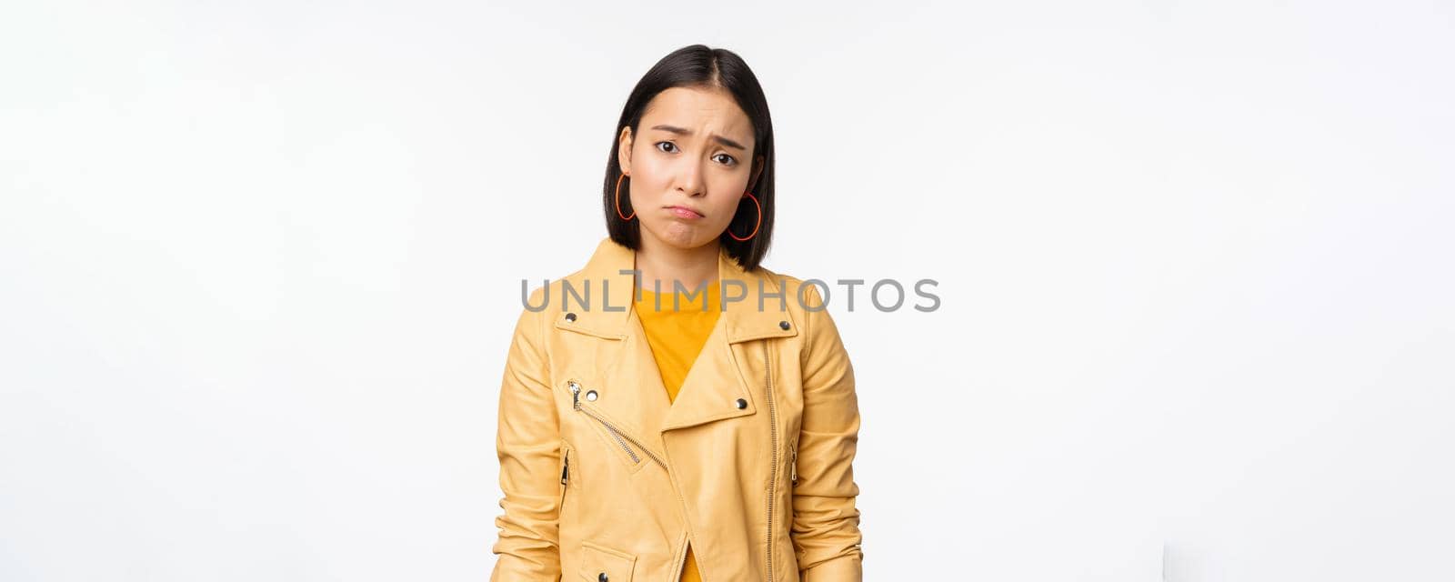 Portrait of sad korean woman sulking, frowning and looking upset, distressed frustrated face expression, standing gloomy against white background.