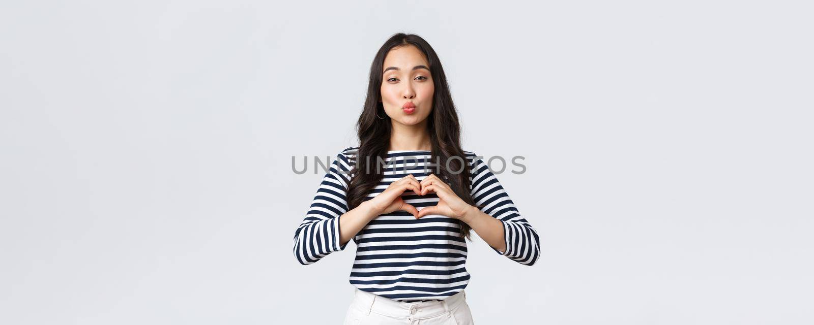 Lifestyle, people emotions and casual concept. Lovely smiling adorable asian woman showing heart sign and smiling, express sympathy or care, sending air kiss at camera, standing white background.