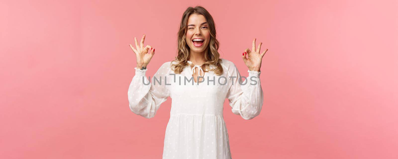 Beauty, fashion and women concept. Portrait of carefree unbothered blond feminine woman in white dress, guarantee quality, wink say no problem, smiling pleased, rate good product, pink background.