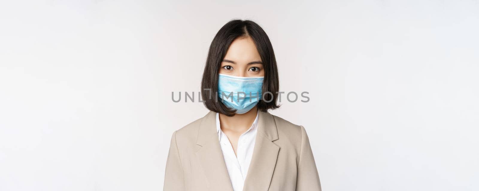 Coronavirus and workplace concept. Portrait of asian office worker, businesswoman in medical face mask looking at camera, white background.