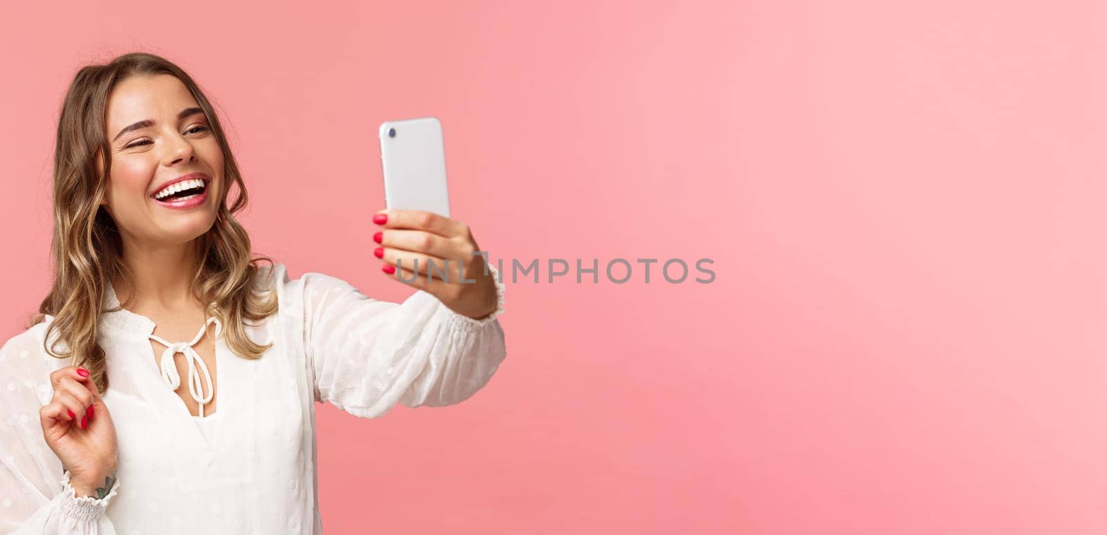 Close-up portrait of cheerful upbeat smiling blond girl, wearing white dress, laughing as record video, calling friend on mobile application, taking photo, selfie on smartphone, pink background.