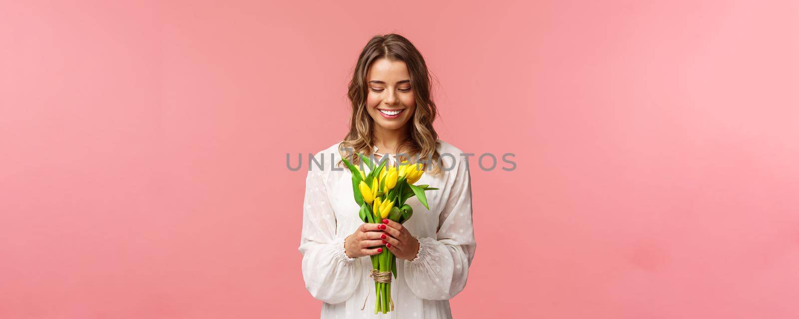 Holidays, beauty and spring concept. Portrait of lovely, romantic blonde girl in white dress, holding yellow tulips, looking at beautiful flowers, smiling and blushing, pink background.