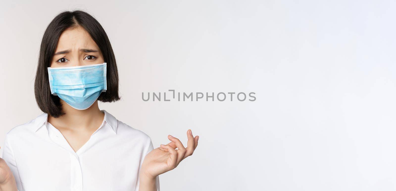 Distressed and miserable young asian woman in face mask, looking up, looking up sad, standing over white background.