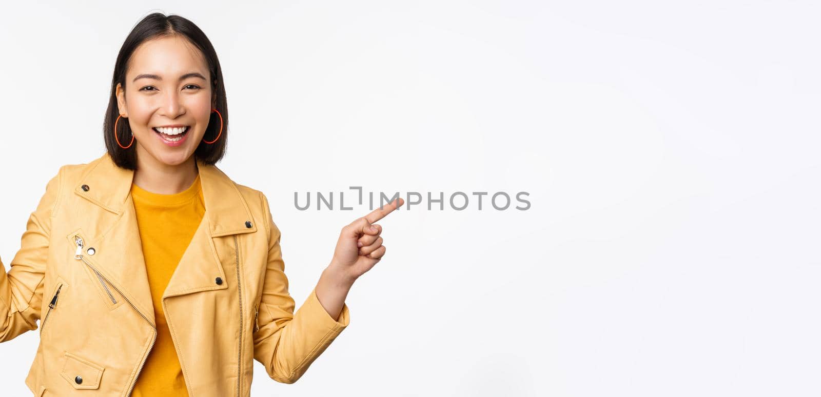 Happy beautiful asian girl pointing fingers left and right, showing banner logo, demonstrating sale, standing in yellow jacket over white background.