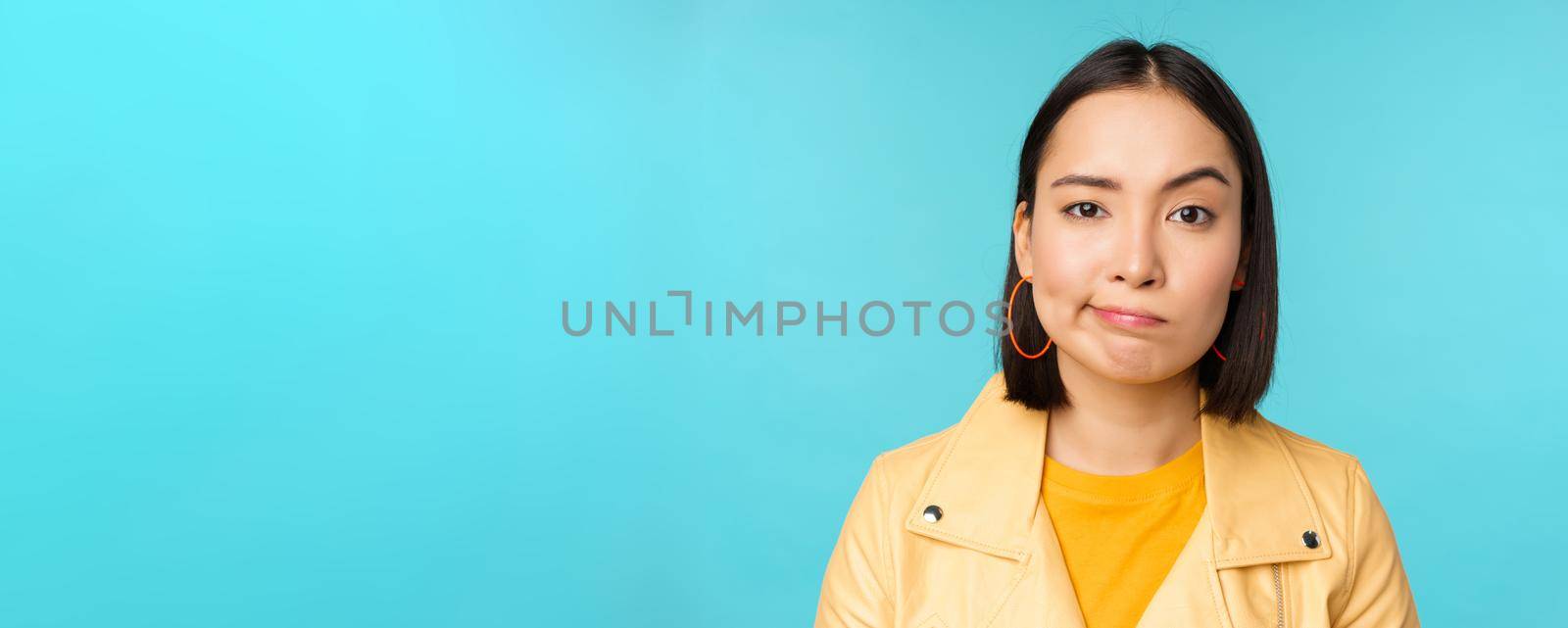 Close up of skeptical asian girl looking disappointed, staring with doubt or disbelief, grimacing at camera, standing over blue background.
